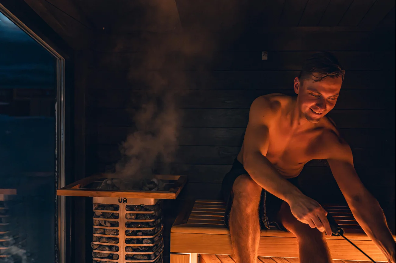 A bather sitting beside a steaming stove in a warmly lit wooden sauna.