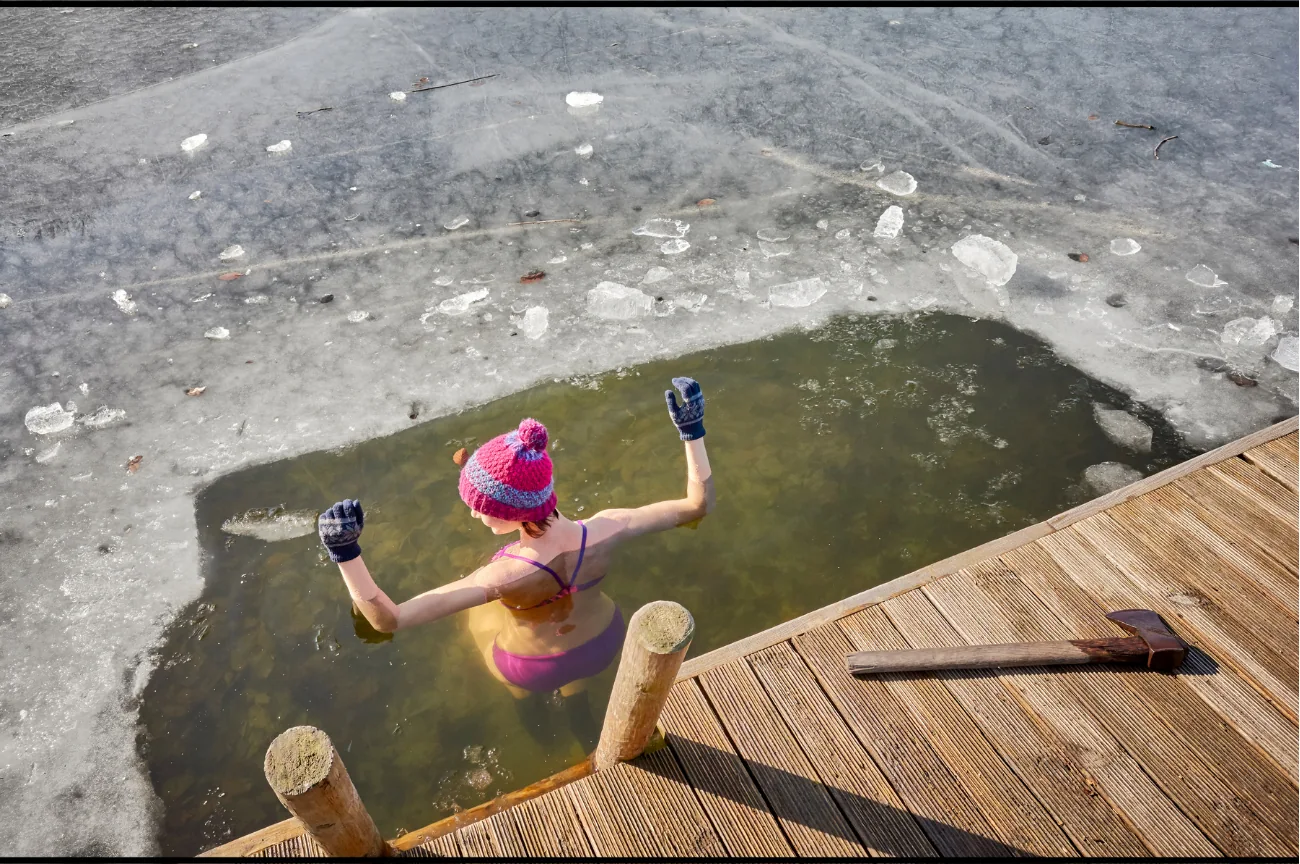 A winter swimmer wearing a bright pink beanie and gloves beside a wooden jetty.